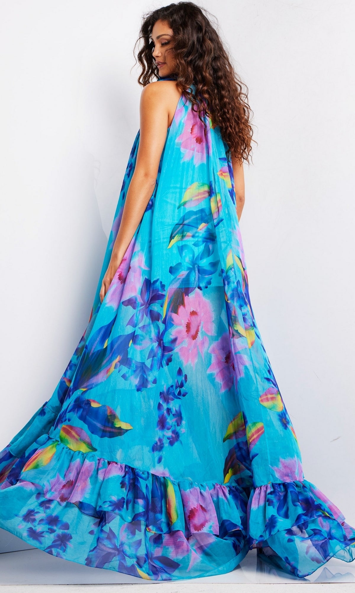A woman with long curly hair wears the Jovani Long Prom Dress 38721, a flowing blue gown with a ruffled hem, standing against a plain white background and looking over her shoulder.